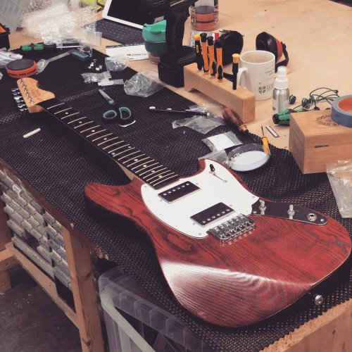 A crimson mustang-style guitar with a wenge freboard sites on a workbench having it's finishing touches added.