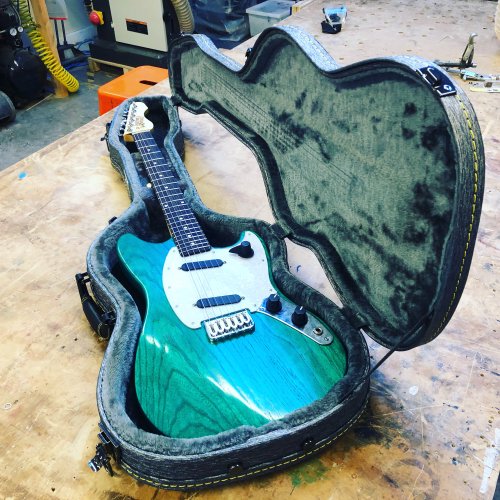 A green mustang-style guitar with a rosewood fretboard sites in an open hardcase upon the workshop bench.