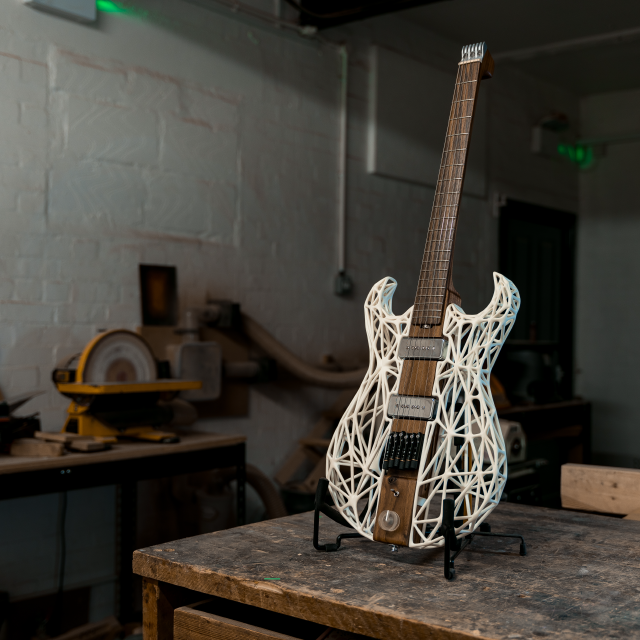 A photo of an unusual looking guitar siting on a stand on a workbench in a moodily lit workshop. The neck and the centre of the body along the neck are made from a single piece of dark walnut wood, whilst the sides of the body are made from a white 3D-printed lattice work. There is no headstock, just a custom aluminium block to anchor the strings, with tuners being built into the bridge.