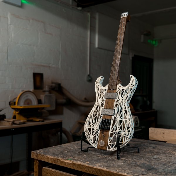 A photo of an unusual looking guitar siting on a stand on a workbench in a moodily lit workshop. The neck and the centre of the body along the neck are made from a single piece of dark walnut wood, whilst the sides of the body are made from a white 3D-printed lattice work. There is no headstock, just a custom aluminium block to anchor the strings, with tuners being built into the bridge.