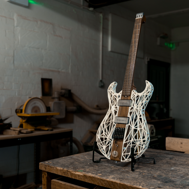 An unusual looking guitar sites on the workbench. The neck and the center of the body along the neck are made from a single piece of dark walnut wood, whilst the sides of the body are made from a white 3D-printed latice work. There is no headstock, just a custom aluminium block to anchor the strings, with tuners being built into the bridge.