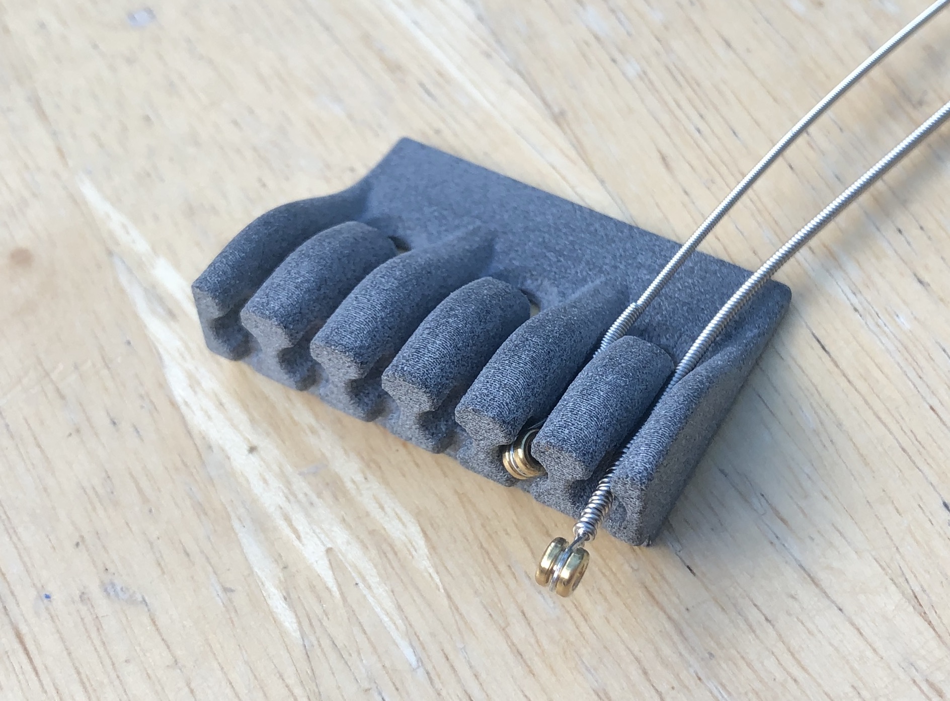 A photo of a 3D-printed plastic part that has 6 channels in it that end in little thimbles. In one channel sits a guitar string with the ball end sat in the thimble. In another channel a string sits over the channel as it's too wide to fit in.