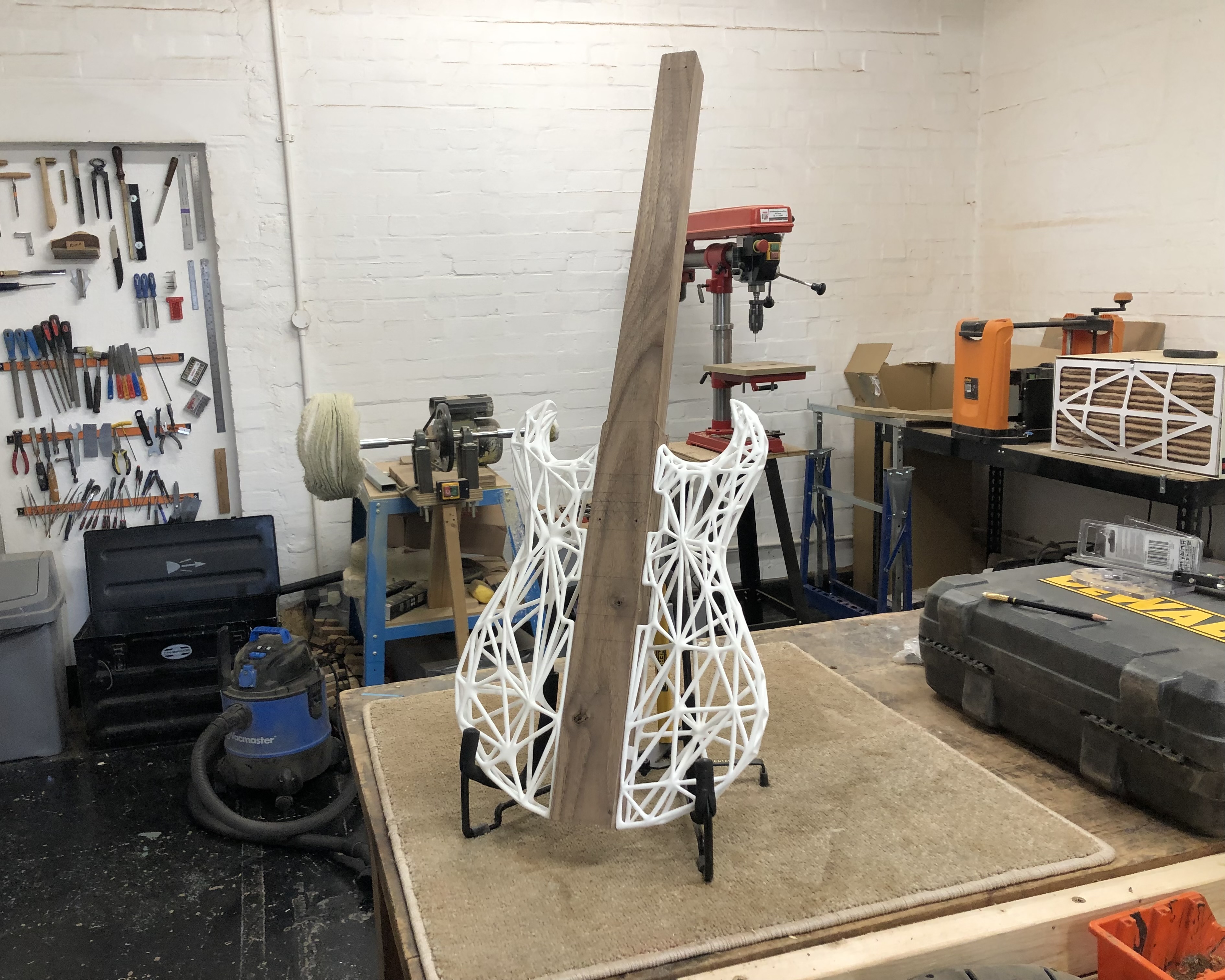 A photo of a weird in progress guitar on stand on a workbench. The centre of the guitar is a plank of walnut that runs the length of the body and neck, and to either side of it the body shape is made from a 3D-printed lattice in white plastic.