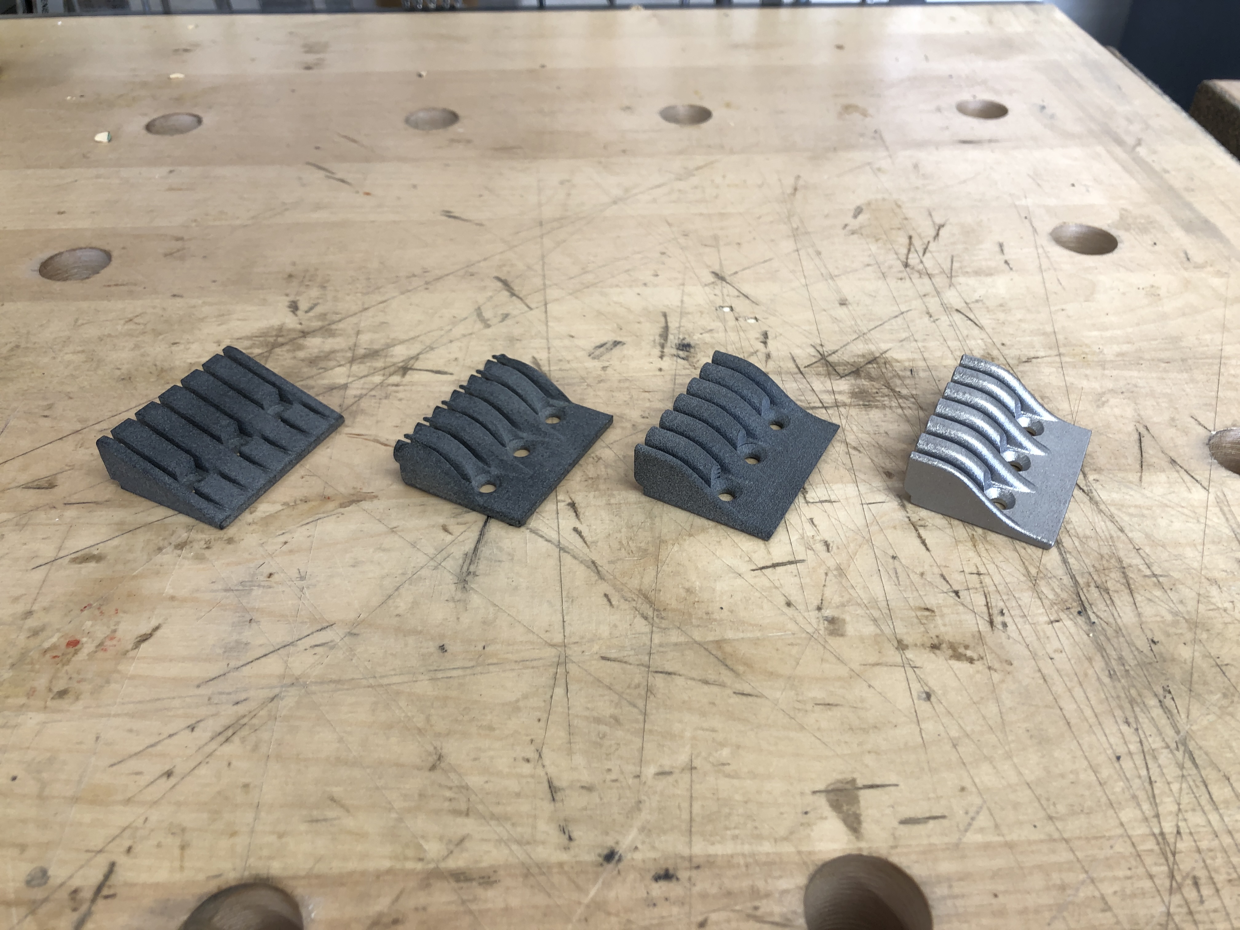 A photo of four wedge shaped parts lined up on a workbench, showing an evolution of the headstock part from left to right. The left most one is very angular and blocky in plastic, the next one is very curved in and bumpy but uses less material and is printed again in plastic, the third is a refinement of the second to have smother lines and is printed in plastic, and the last one is the same as the third but printed in aluminium.