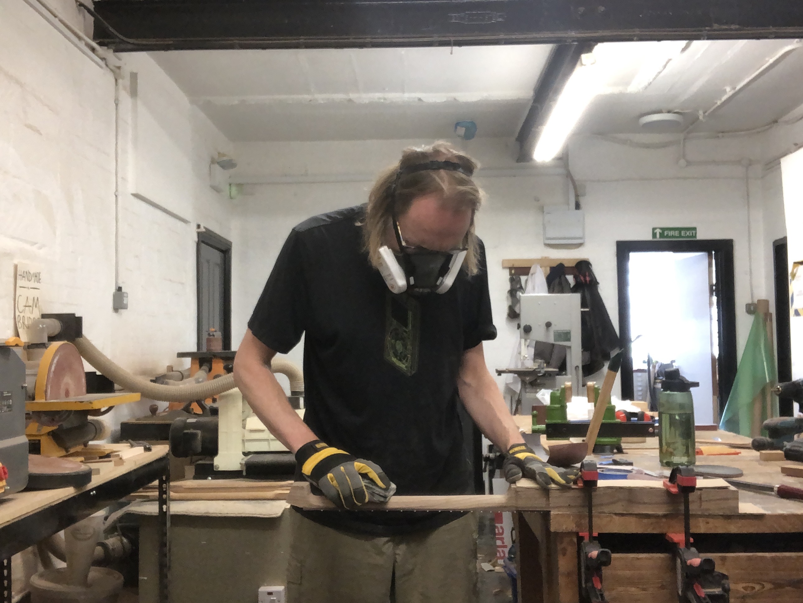 A photo of me in the workshop with the neck section of Älgen clamped to the edge of a workbench, and I'm carving the back of the neck, wearing my respirator.