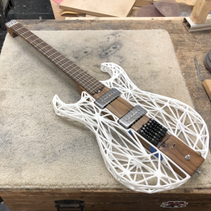 An unusual looking guitar sites on the workbench. The neck and the center of the body along the neck are made from a single piece of dark walnut wood, whilst the sides of the body are made from a white 3D-printed latice work. There is no headstock, just a custom aluminium block to anchor the strings, with tuners being built into the bridge.