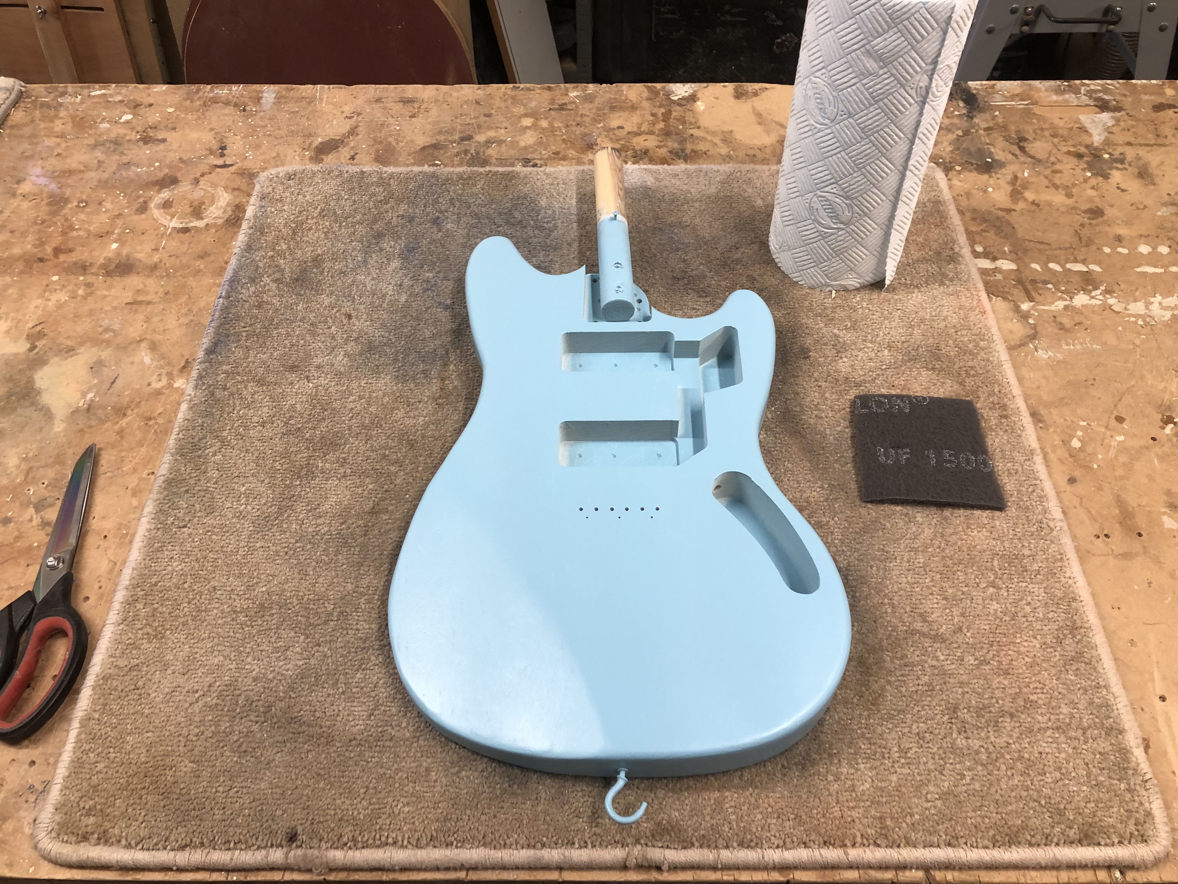 A photo of an offset-style guitar body on the workbench, painted a pale blue colour. Next to it sits a sanding pad, some scissors, and a roll of kitchen-roll.