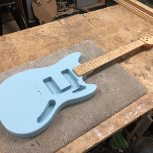 On the workbench sits a work-in-progress guitar, with just the painted-body and neck in position. The body is in the style of a Fender Mustang offset guitar and is painted a pale blue, and the neck is made from birds-eye maple and finished in oil.