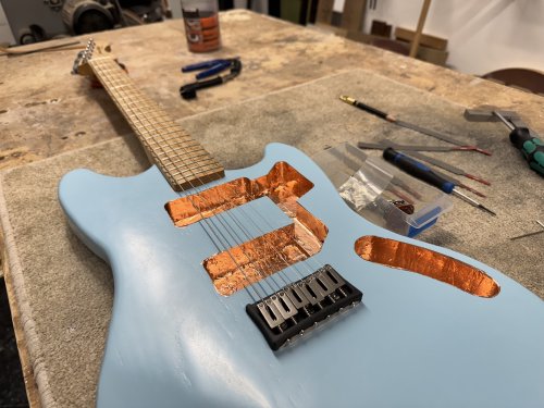 On the workbench sits a work-in-progress offset solid-body electric guitar, with a painted light blue body, and an all maple neck. There are now electronics, but you can see copper tape in all the cavities in the body for them. There is a carbon-fibre printed bridge and strings are installed.
