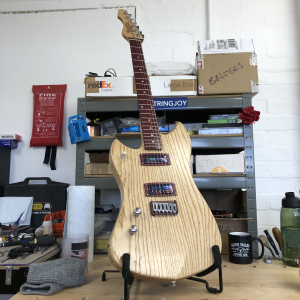 A left handed smaller stature guitar sits in a stand on the workbench. The body is a somewhat pointy in style, with natural ash wood showing through an oil finish. The fretboard is purpleheart wood on birds-eye maple. The pickup rings and bridge are also made from purpleheart wood.
