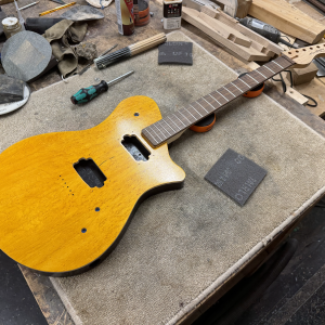 An in-progress guitar shot, showing a body with birds-eye maple cap and various bits of hardware like bridge and pickup rings laid on the body, and a maple neck with rose-wood fretboard sat in position.