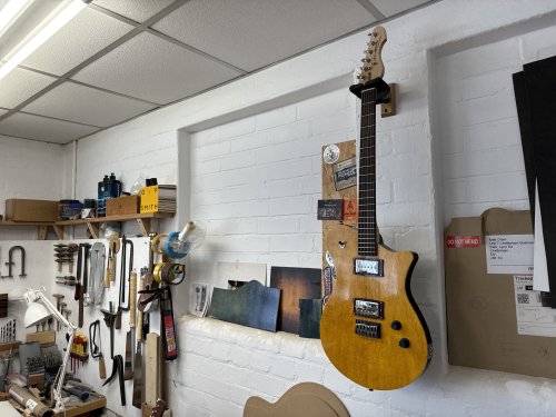 A photo of a completed guitar hung on the wall in the workshop, showing a body with birds-eye maple cap stained bright yellow, and black sides, with a maple neck and a rosewood fretboard. It has a chrome covered P90 in the neck position and a chrome covered humbucker in the bridge position.
